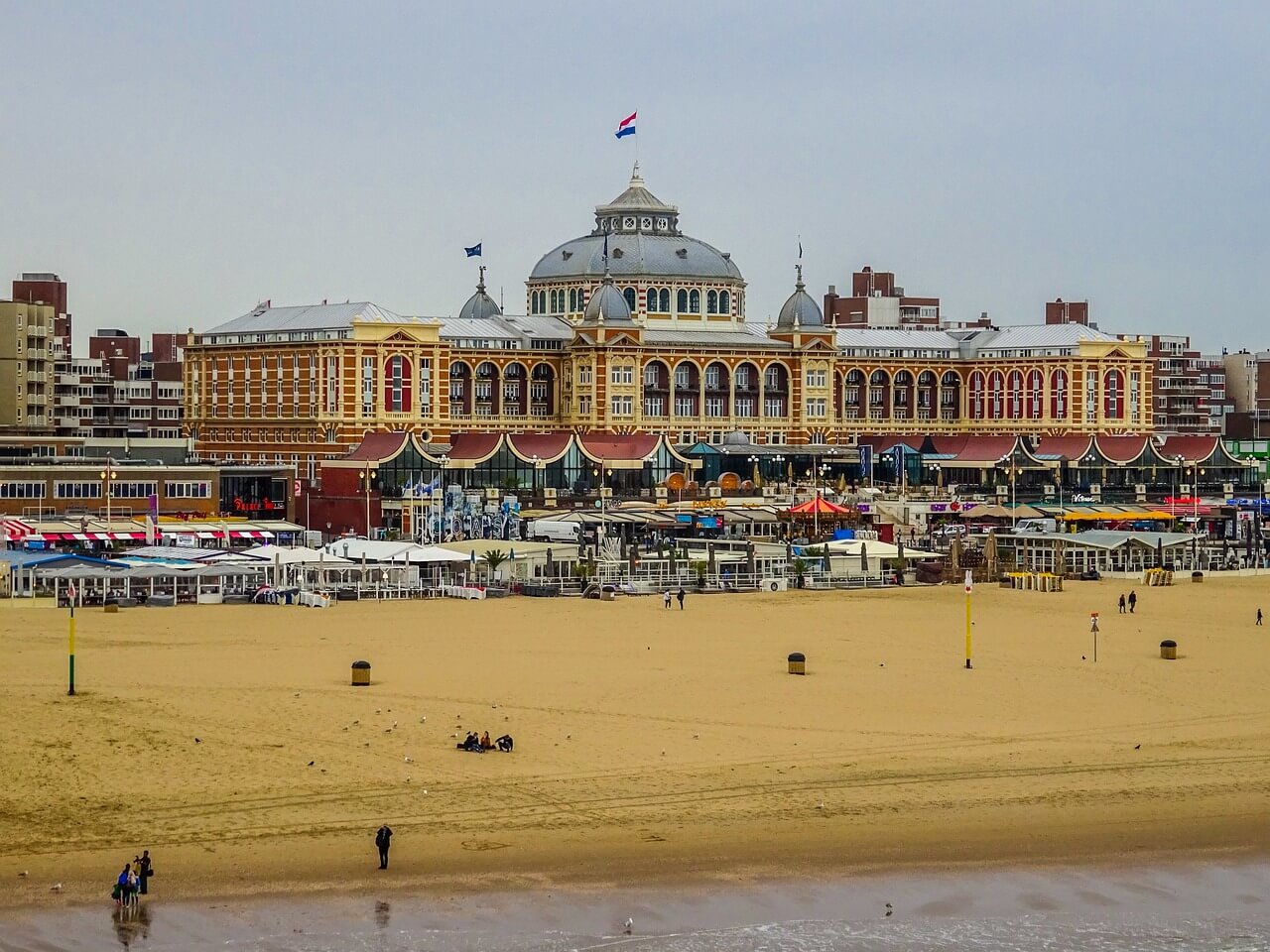 netherlands-6514965_1280 "Beachfront view of a grand building with a domed roof and multiple flags, featuring cafes and restaurants along the shoreline, under a clear blue sky.